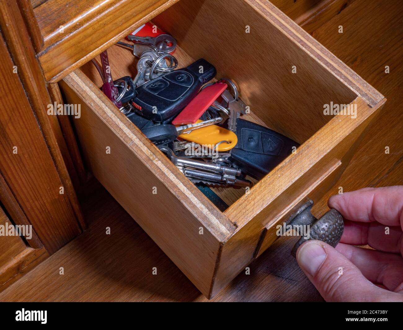 Close overhead shot of man’s fingers on the knob of a small open drawer in an old pine unit, holding an assortment of vehicle  door and ignition keys. Stock Photo