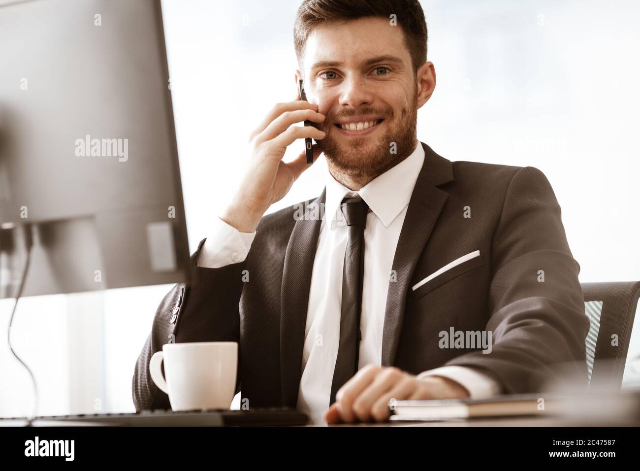 Business concept. Young businessman sitting at the office table happy talking on a cell phone getting good news about his work. Man in suit indoors on Stock Photo