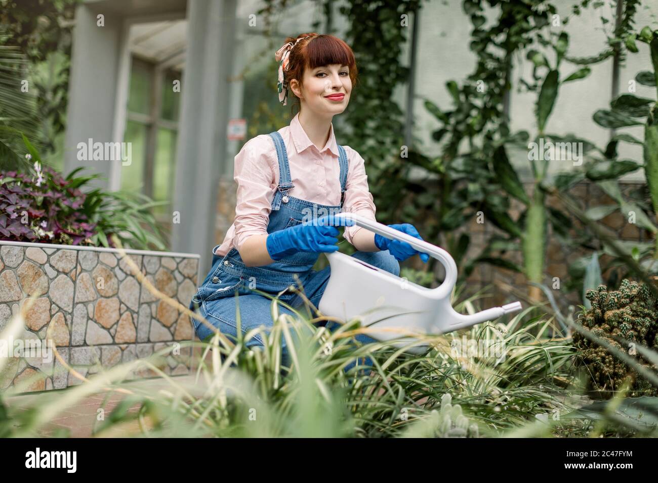 Young beautiful woman in protective gloves and overalls, watering plants in her home garden or in a greenhouse, smiling and looking at the camera Stock Photo