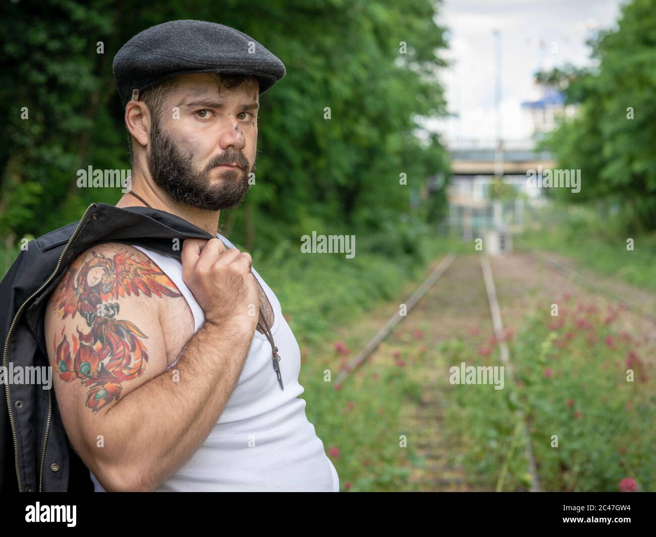 Young french handsome bearded man, with tattoos and charcoal stains on his skin, taken outside on a disused railroad track with flowers using natural Stock Photo