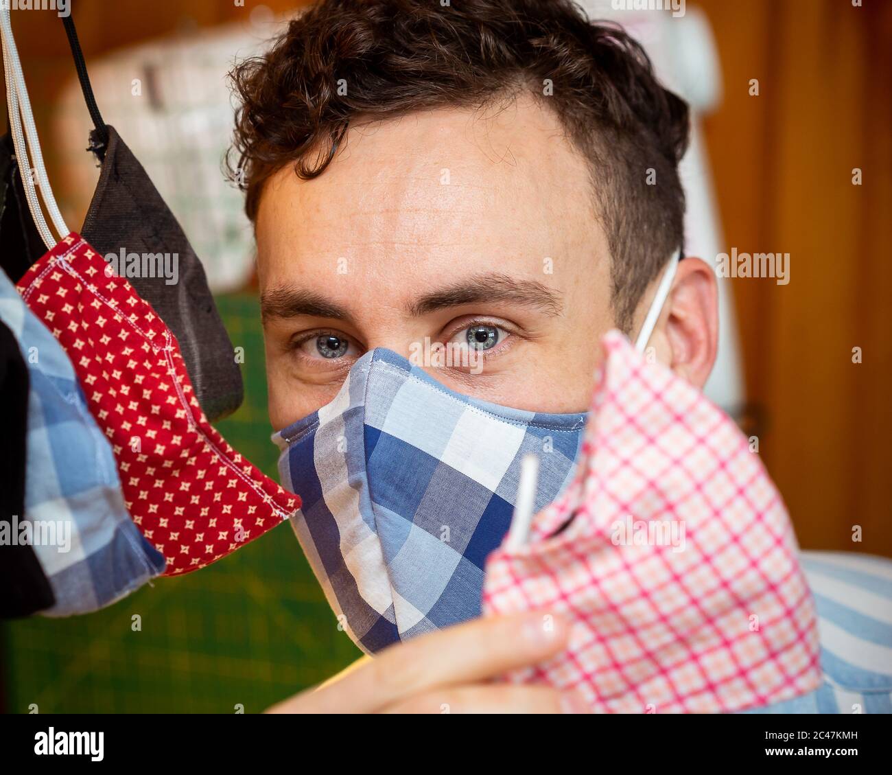 Young handsome male mask maker displaying his deluxe handmade masks blue eyes dark hair good bone structure pink plaid red stars black big blue checks Stock Photo