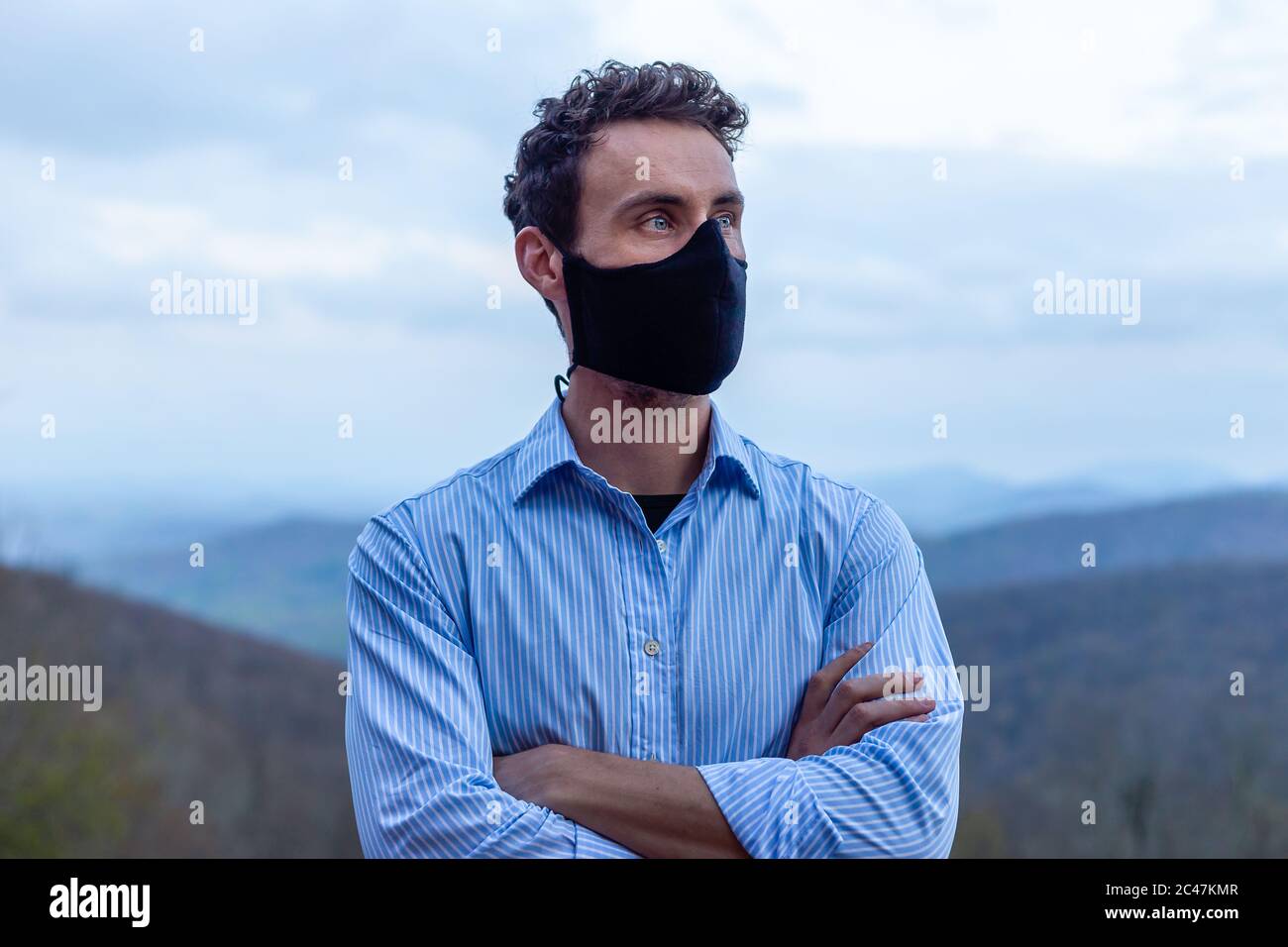 Young man wearing black mask looking confidently into the future blue button up shirt blue eyes dark brown hair good bone structure crossed arms rich Stock Photo
