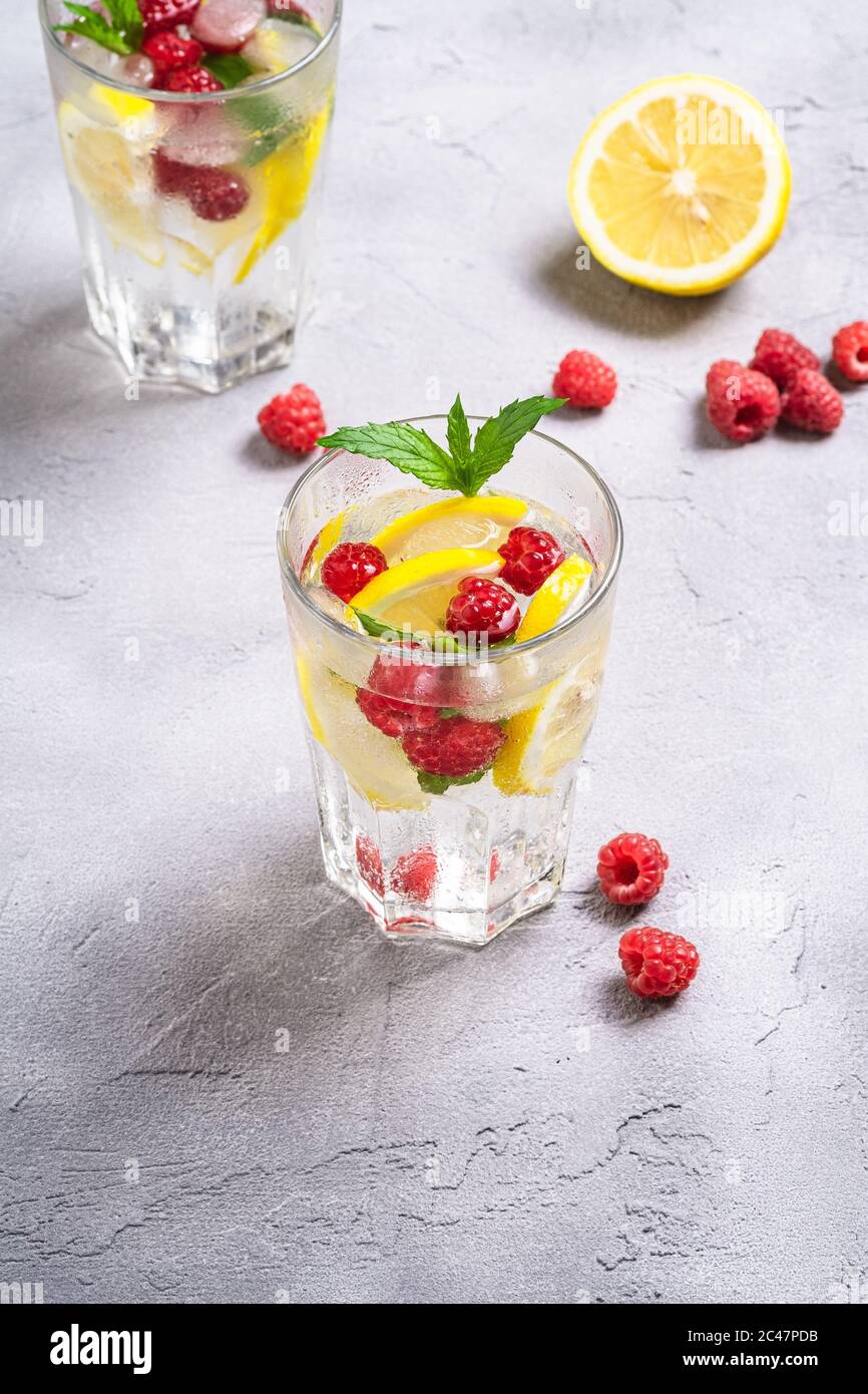 Fresh cold ice water with lemon, raspberry fruits and mint leaf in two faceted glass on stone concrete background, summer diet beverage, angle view Stock Photo