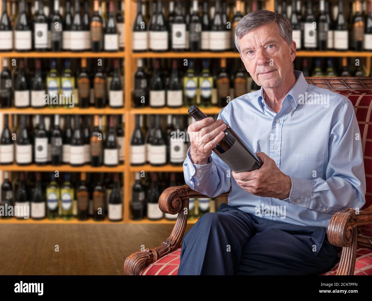 Friendly senior retired man examining a wine bottle label while seated in front of shelves in his wine cellar Stock Photo