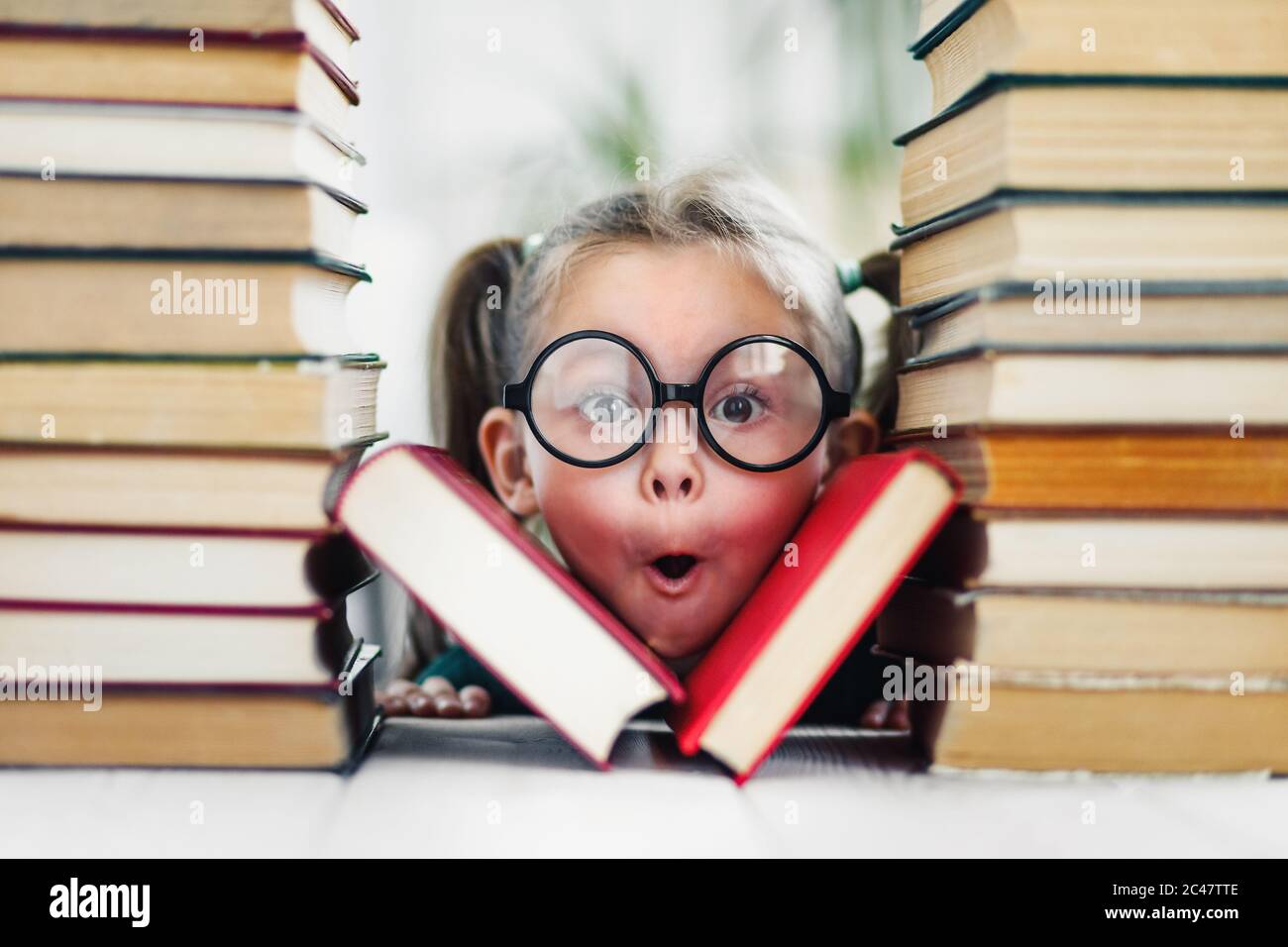 Preschool age girl with wondering face in big funny glasses among a pile of books Stock Photo