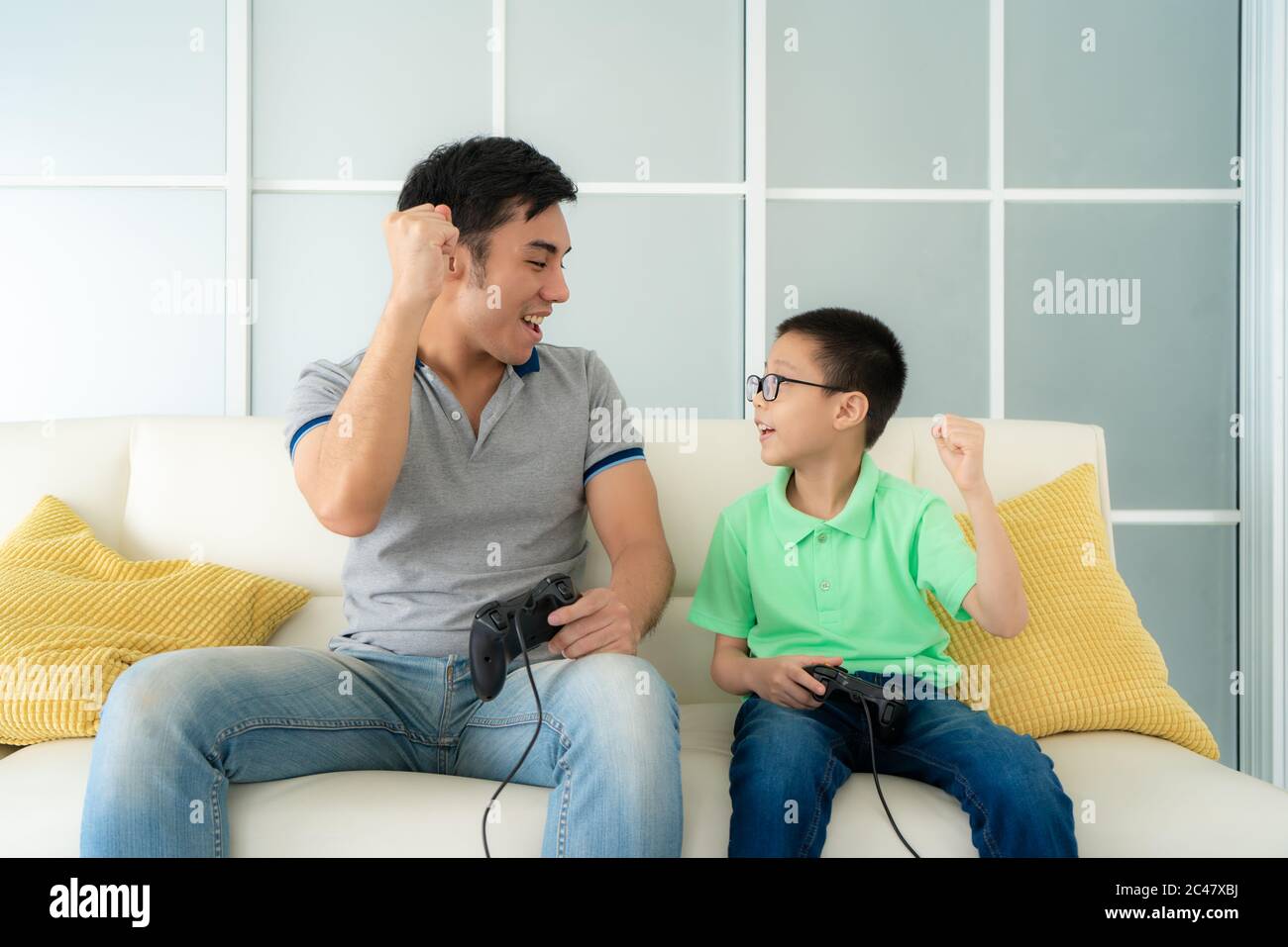 Asian family father and his son to playing video games with joysticks and arm raised to winning in game while sitting in sofa in living room at home, Stock Photo