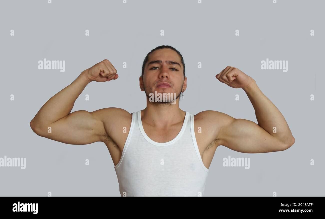 Young hispanic man with hair pulled up bun in white sleeveless t-shirt showing muscles of his arms looking towards camera on light gray background Stock Photo