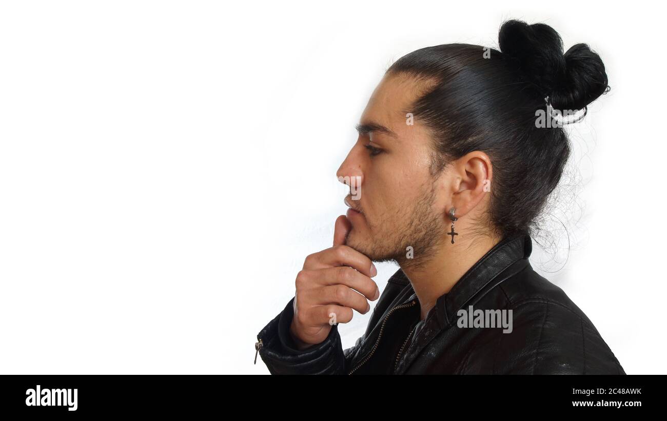 Young hispanic man with gathered hair done bow wearing black t-shirt and black leather jacket, with his hand on jaw with thoughtful attitude, seen in Stock Photo