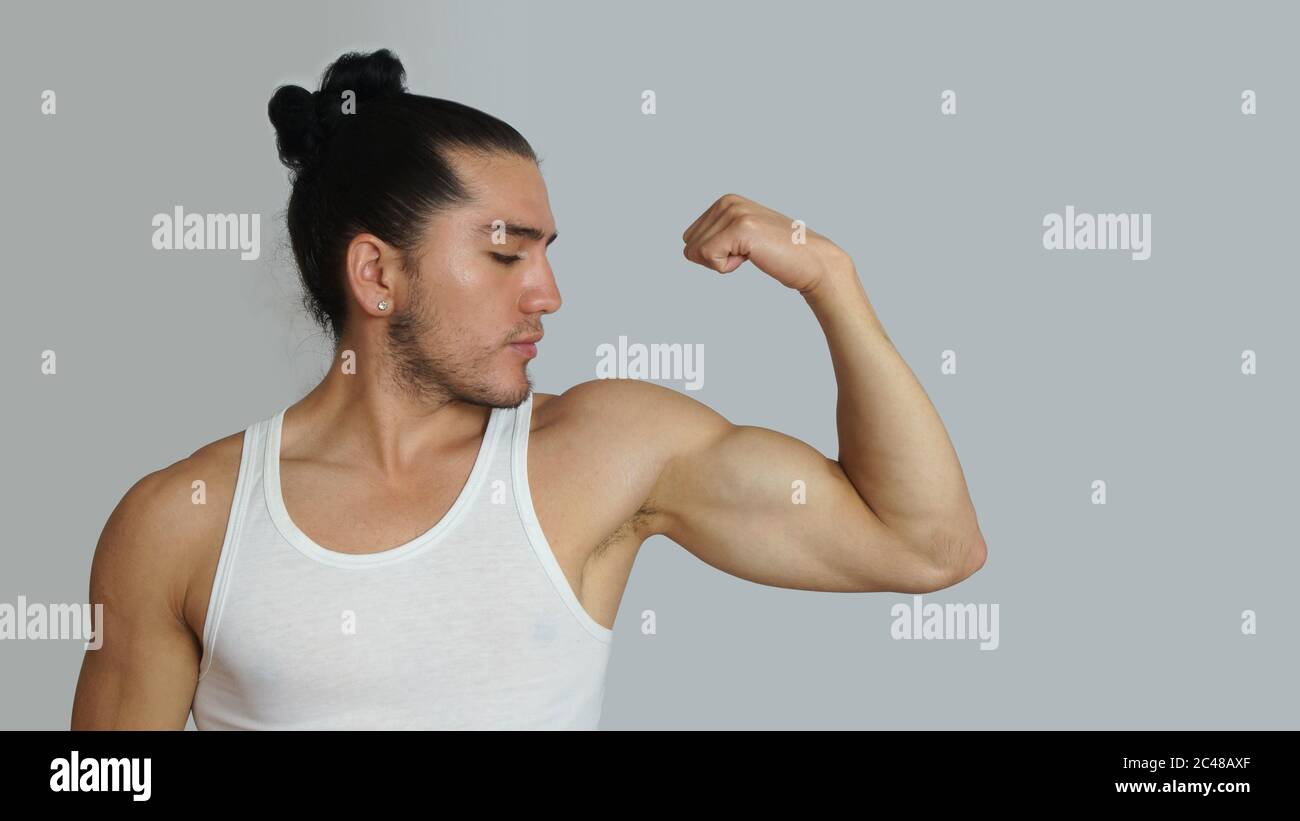 Young hispanic man with hair pulled up bun in white sleeveless t-shirt showing muscles of his arm, looking towards his arm on light gray background Stock Photo