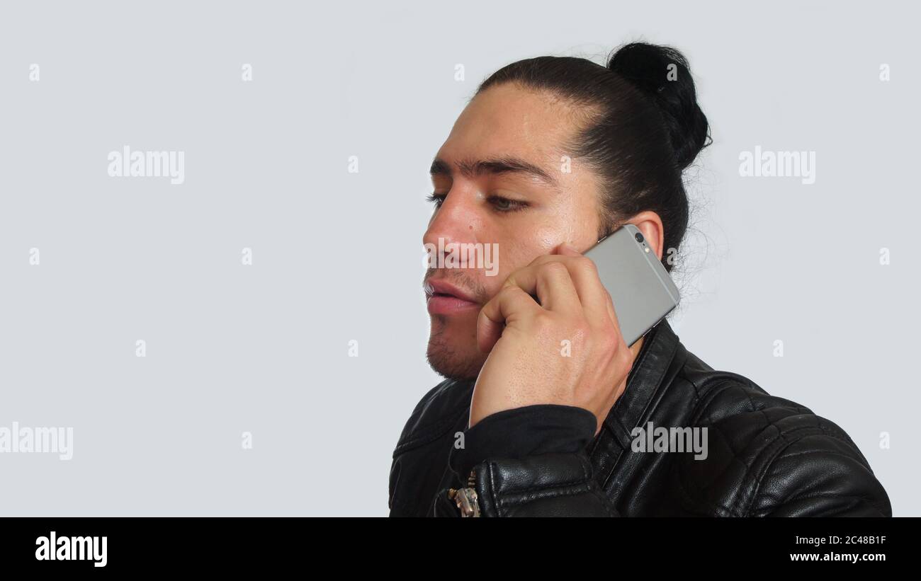 Young hispanic man with gathered hair done bow in black dress and black leather jacket, using mobile phone, seen in profile on white background Stock Photo