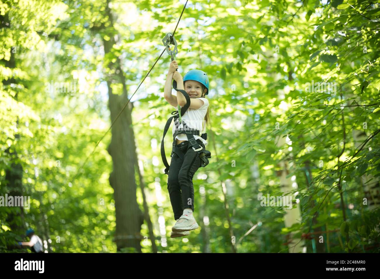 A little girl hanging on the insurance cable of her belt - rope bridge entertainment attraction. Mid shot Stock Photo