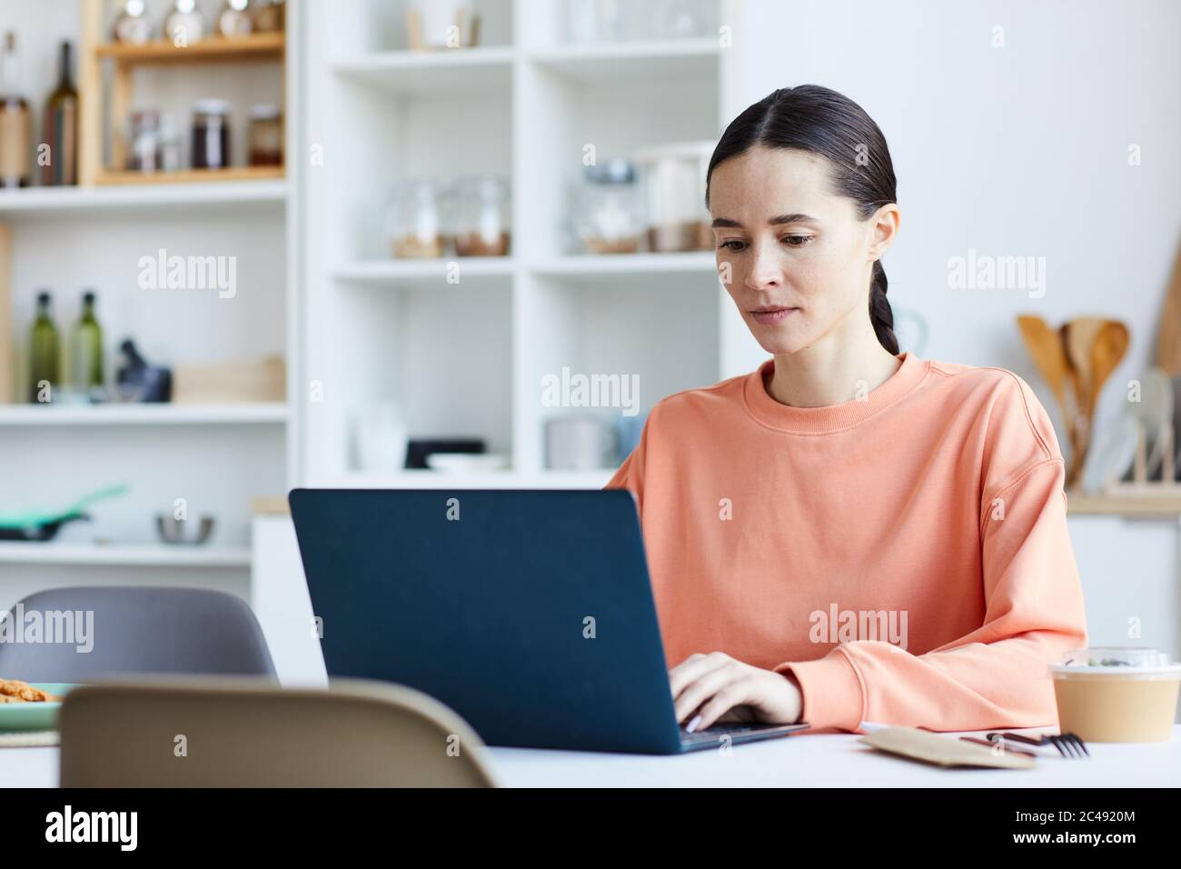 Young woman using laptop computer at her work while sitting at the table in the kitchen Stock Photo