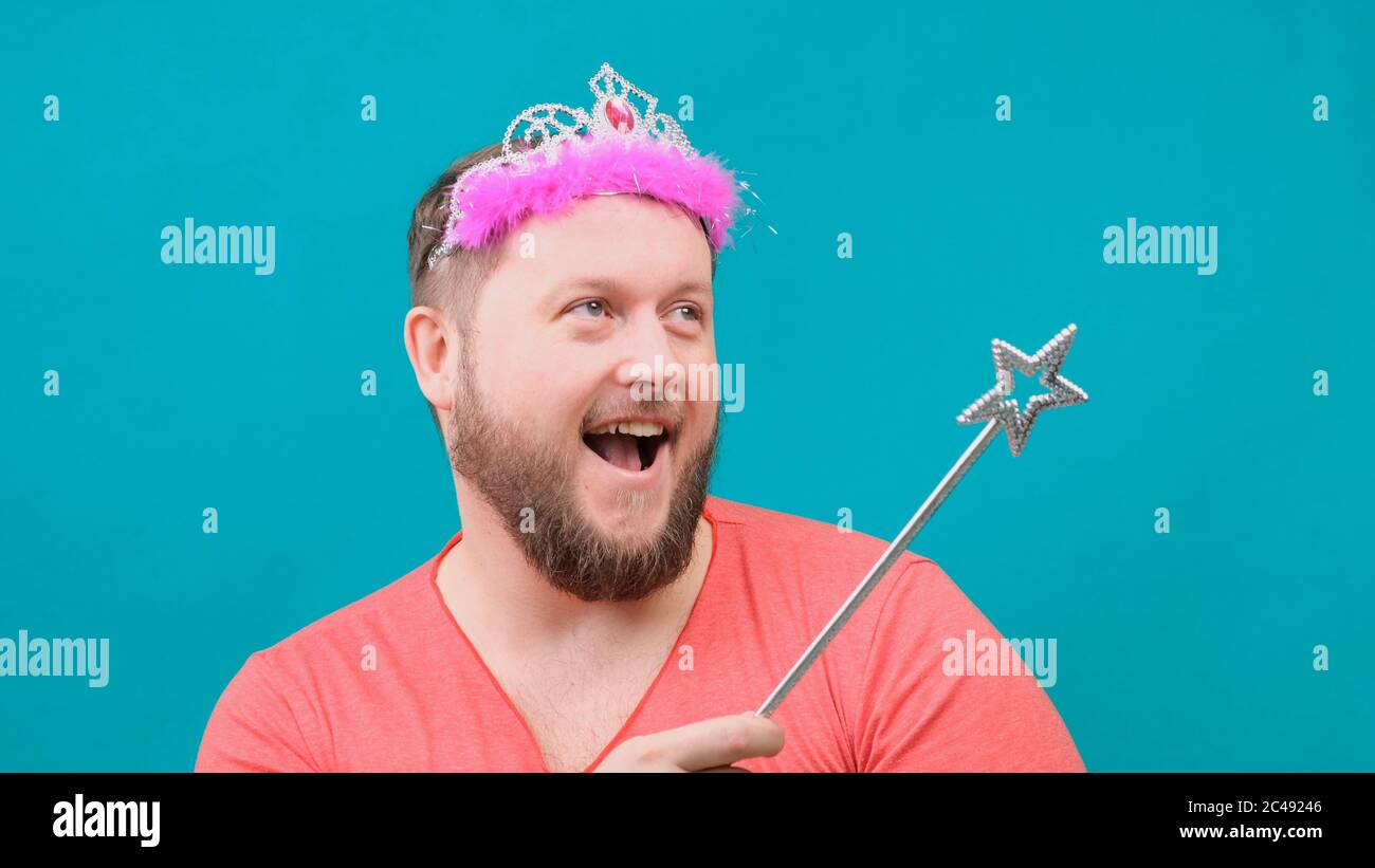 funny bearded freaky man in a pink T-shirt with a deadema on his head is dreaming with a magic wand in his hand. A funny wizard joke to make and Stock Photo