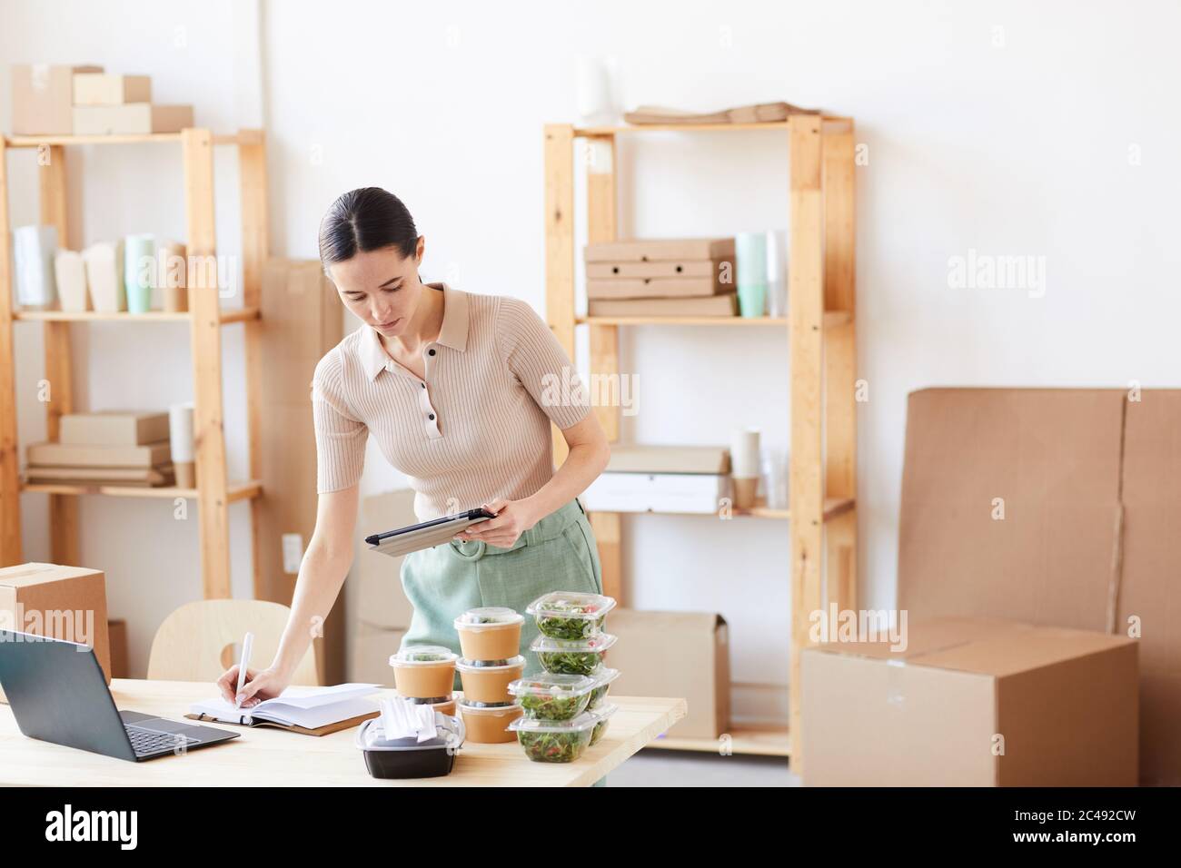 Young woman making notes in note pad and using digital tablet she making a delivery at office Stock Photo
