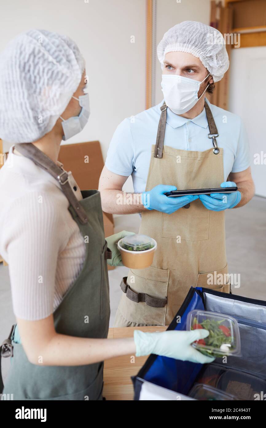 Young man in protective mask using digital tablet and talking to his colleague while they packing food for delivery Stock Photo