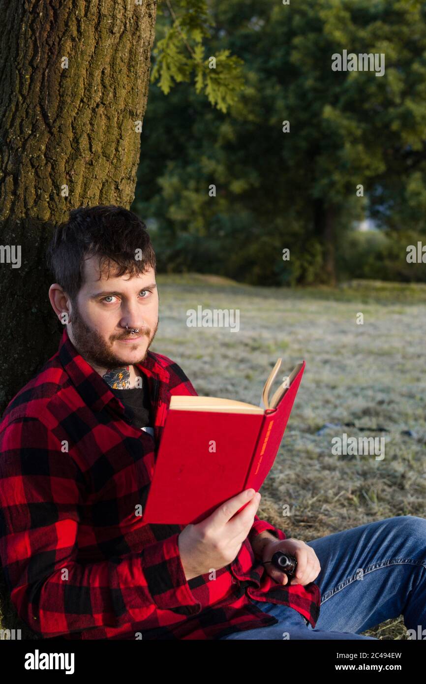Closeup shot of a young male in a red suit reading a book sitting and leaning on a tree Stock Photo