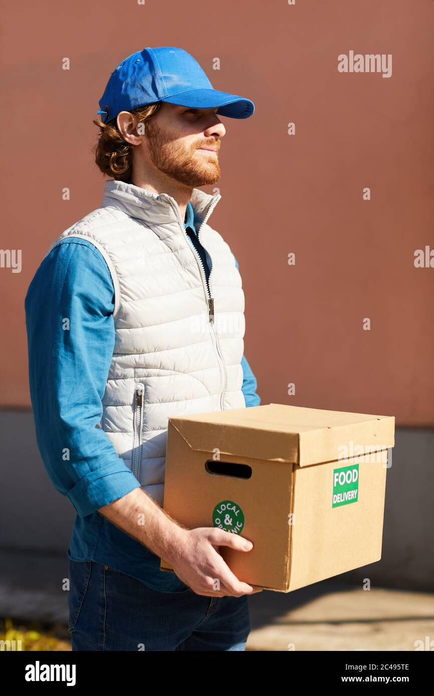 Young man in uniform carrying cardboard box with food outdoors he delivering the food Stock Photo
