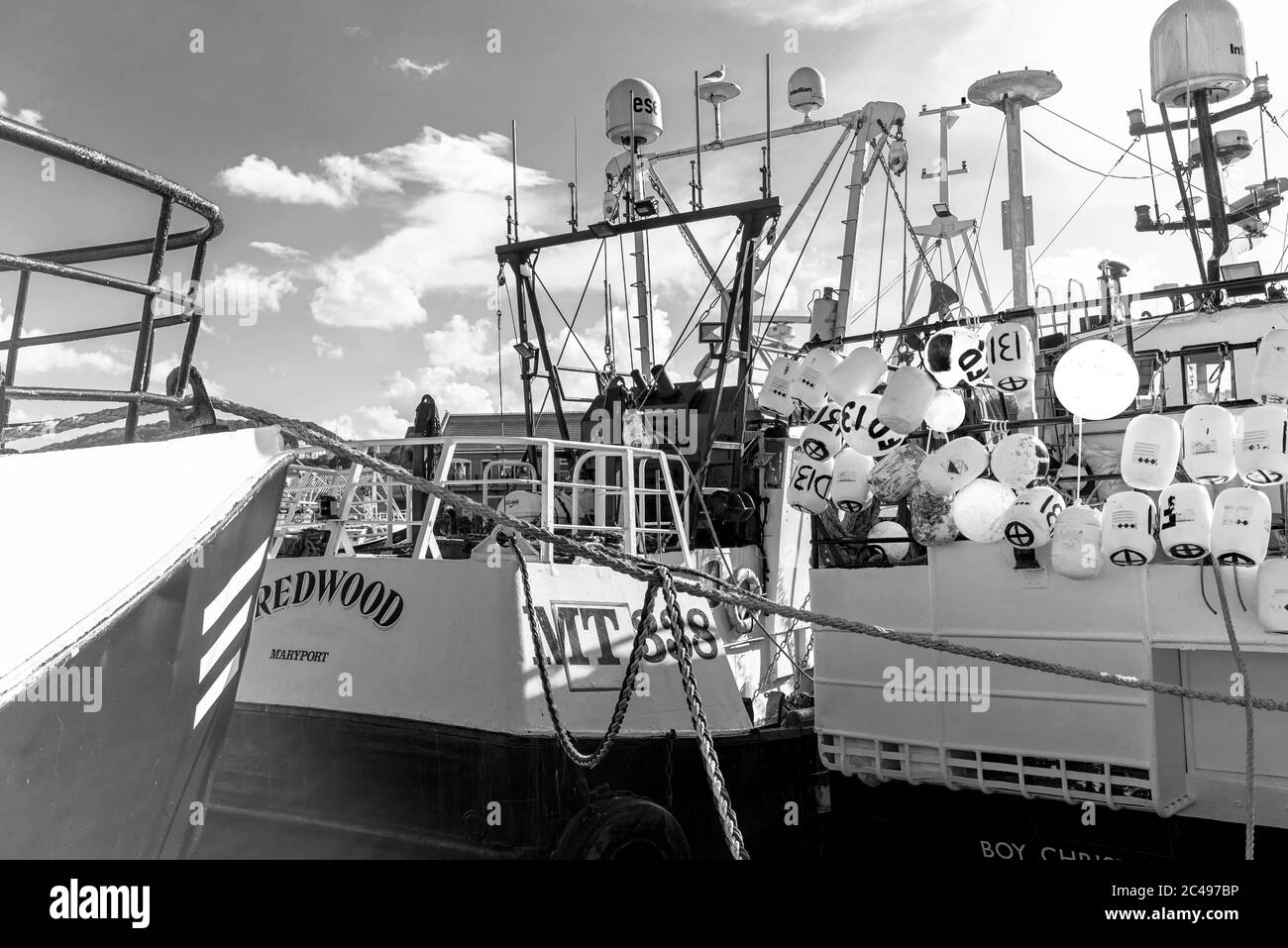 The sterns of two fishing boats moored in harbour.  One is festooned with marker buoys and the bow of another boat is in the foreground. Stock Photo