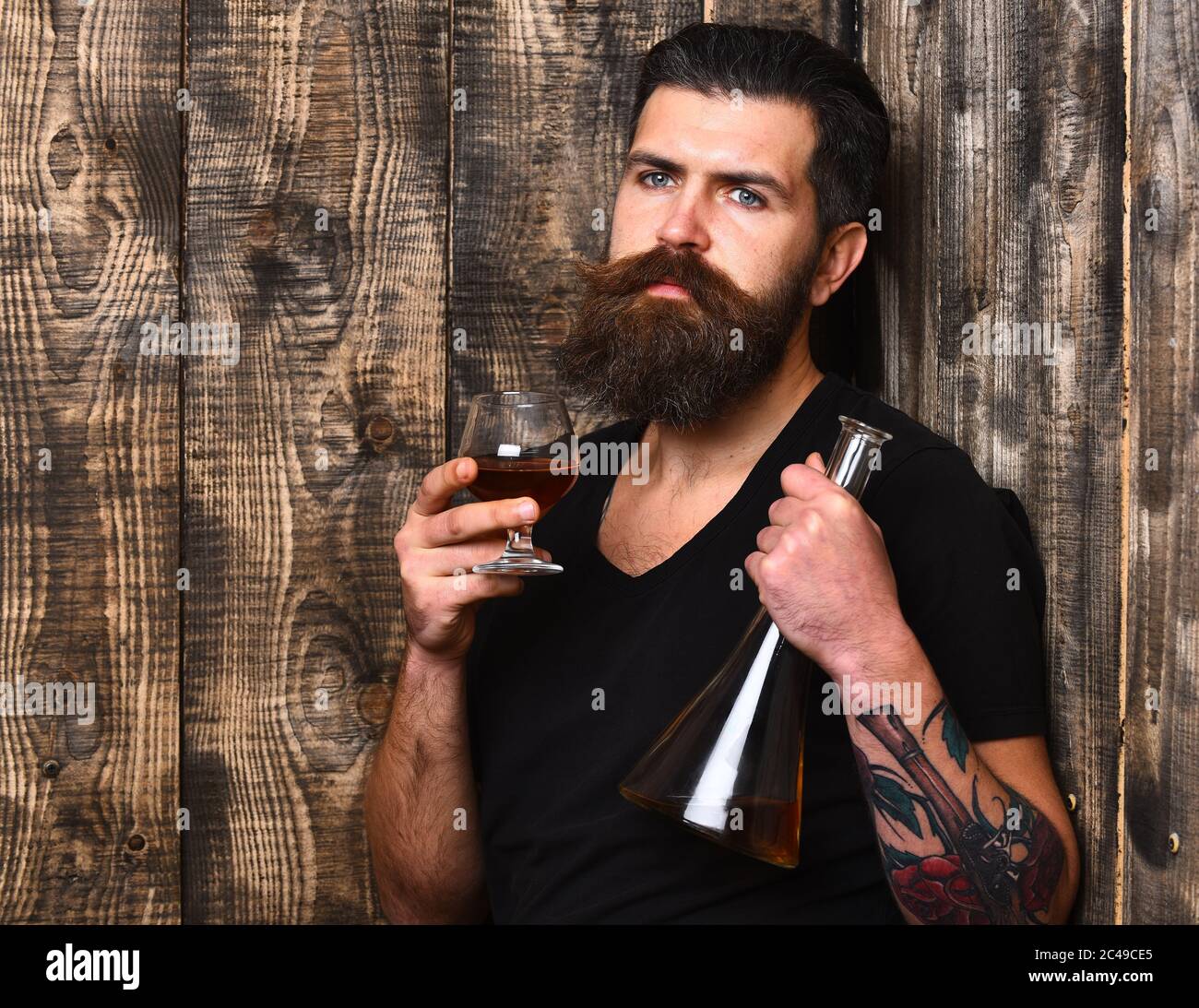 Man with beard and mustache holds alcoholic beverage on wooden wall background. Guy with glass and bottle of cognac. Service and catering concept. Macho with thoughtful face drinks brandy or whiskey. Stock Photo