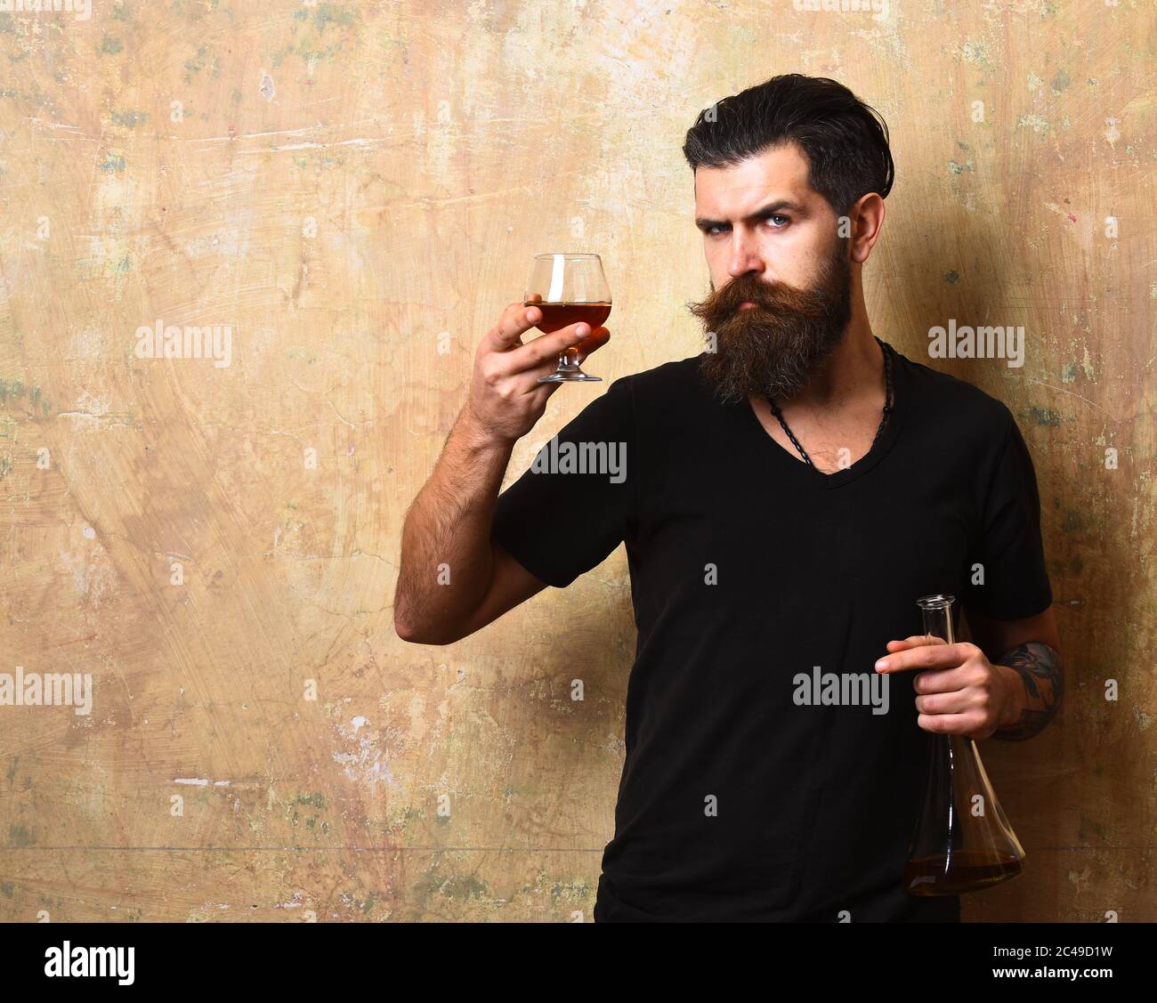 Macho with serious face drinks brandy or whiskey. Man with beard and mustache holds alcoholic beverage on beige background, copy space. Service and tasting concept. Guy with glass and bottle of cognac Stock Photo