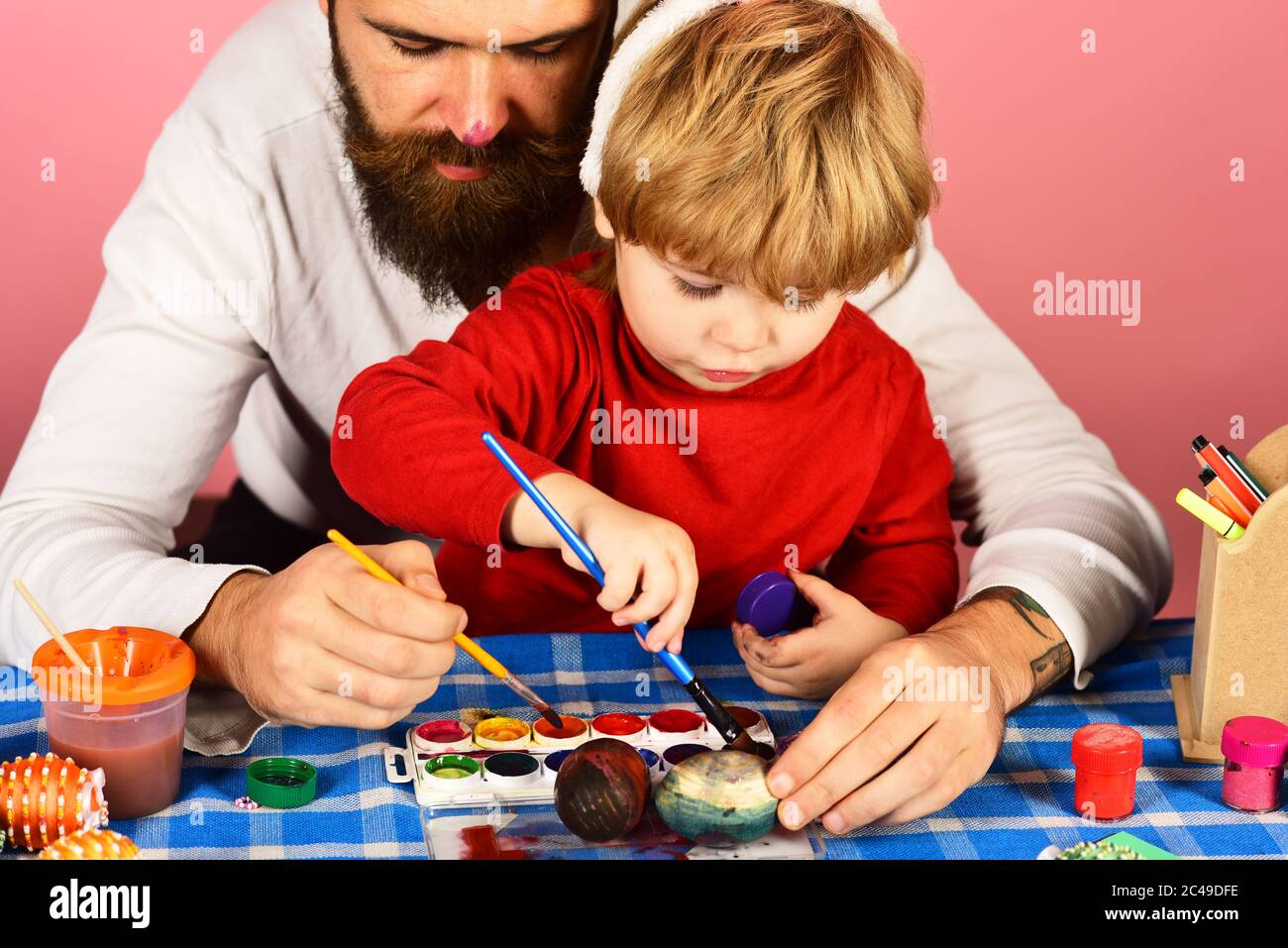 Easter celebration and joy concept. Man with beard and little boy painting eggs for Easter on pink background. Father and son preparing for holiday. Dad with painted nose make decorations Stock Photo