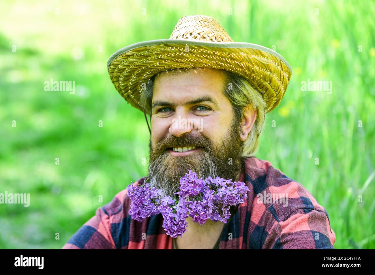 Rustic man with beard happy face enjoy life in ecological environment.  Hipster with lilac flowers looks happy. Bearded man with lilac in beard  green grass background. Eco friendly lifestyle concept Stock Photo 