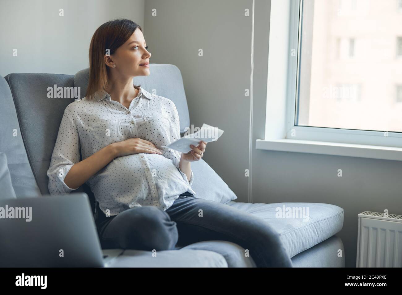 Dreamy lady with gadget by her side sitting on the sofa alone Stock Photo