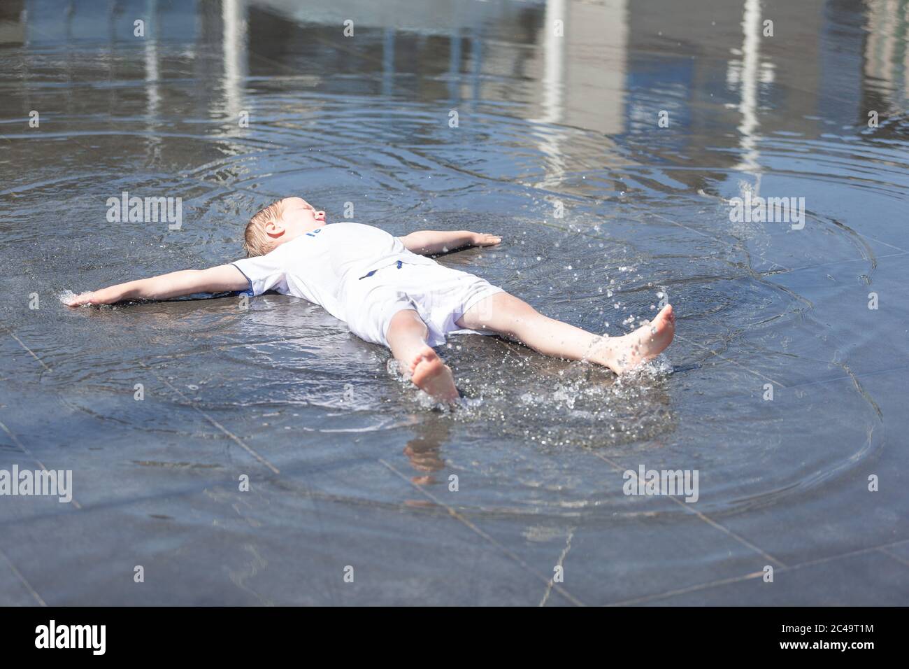 Little boy lying in shallow water fountains on a hot day, UK Stock Photo