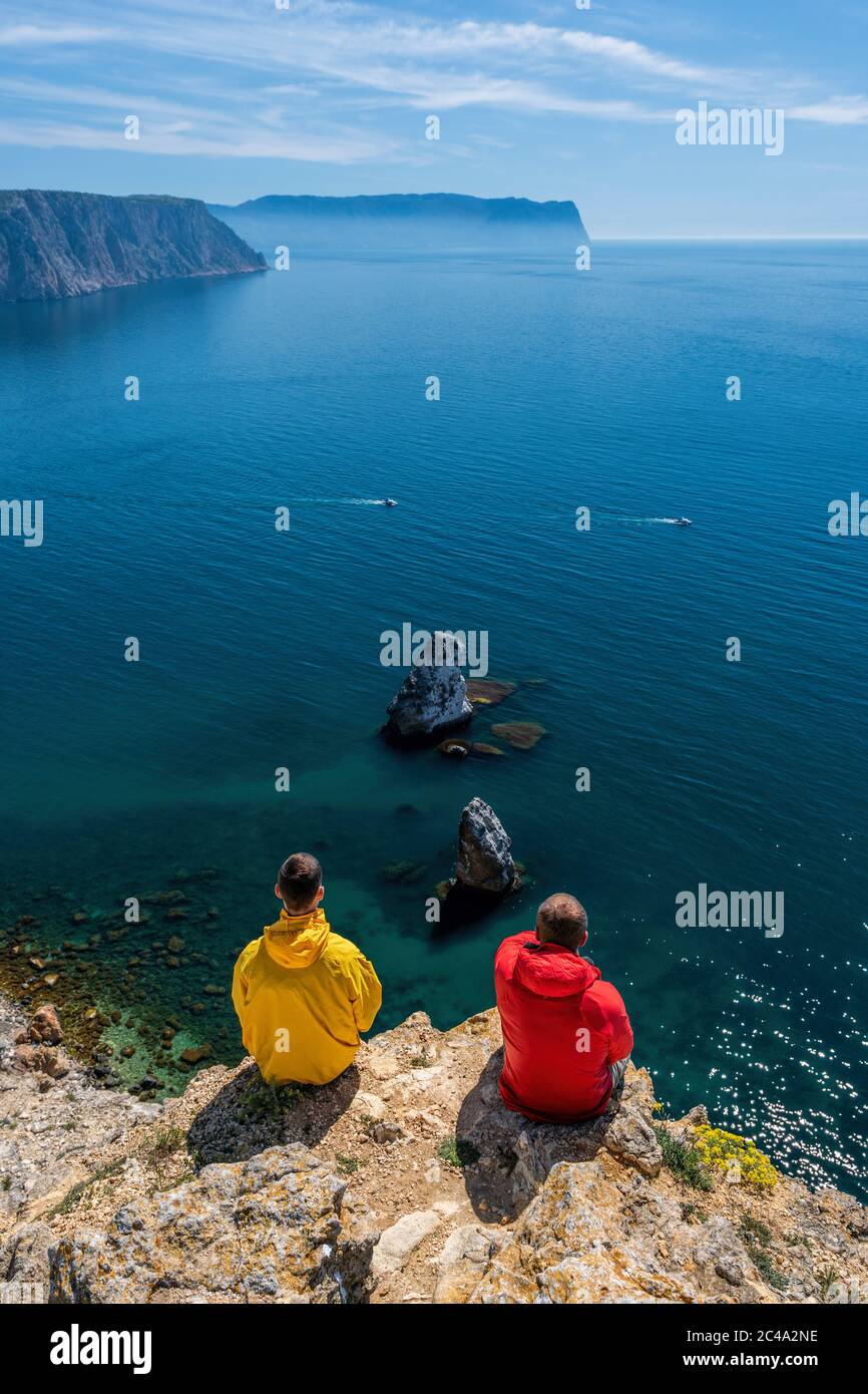 Two caucasian young men travelers in yellow and red jackets sitting high above sea, on the background of coastal cliffs, calm clear blue sea, rocks Stock Photo
