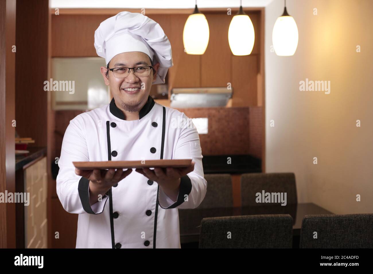 Portrait of Asian male chef looks happy and proud presenting something on his empty wooden plate, copy space meal menu concept Stock Photo