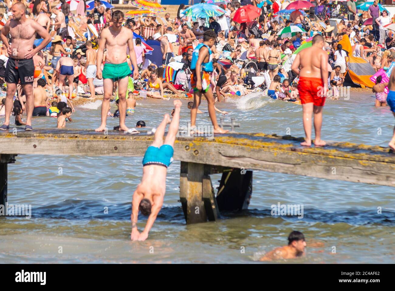 Southend on Sea, Essex, UK. 25th Jun, 2020. With the forecast record temperatures for 2020 people are heading to the seafront to cool down, despite the COVID-19 Coronavirus advice. In Southend on Sea the shore of the Thames Estuary is packed, with people swimming and jumping into the sea. White Caucasian male diving into the sea from a wooden pier with packed beach beyond Stock Photo