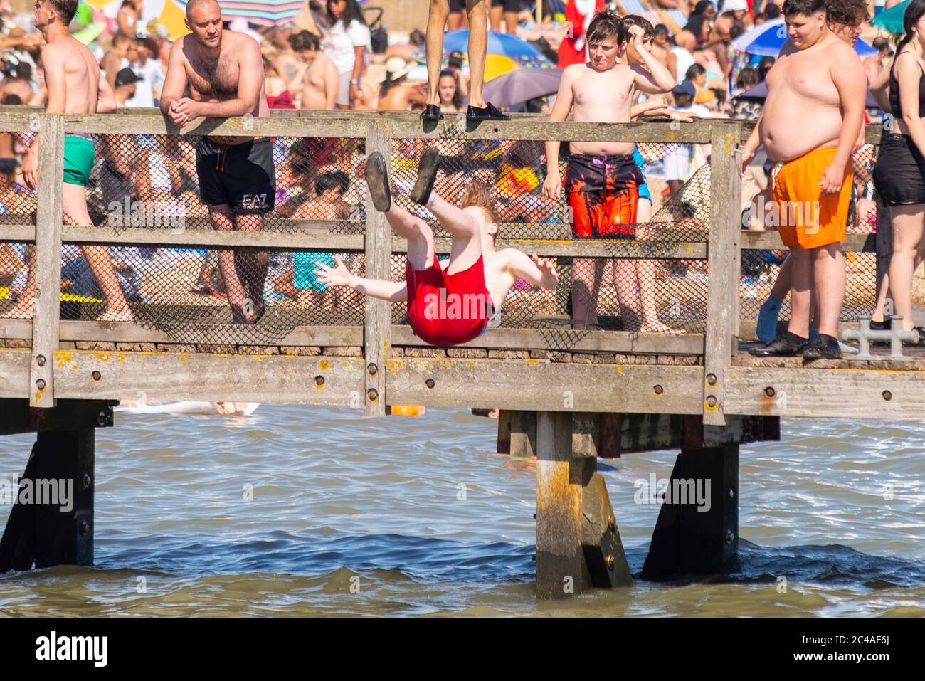 Southend on Sea, Essex, UK. 25th Jun, 2020. With the forecast record temperatures for 2020 people are heading to the seafront to cool down, despite the COVID-19 Coronavirus advice. In Southend on Sea the shore of the Thames Estuary is packed, with people swimming and jumping into the sea. White Caucasian male jumping into the sea from a wooden pier with packed beach beyond Stock Photo