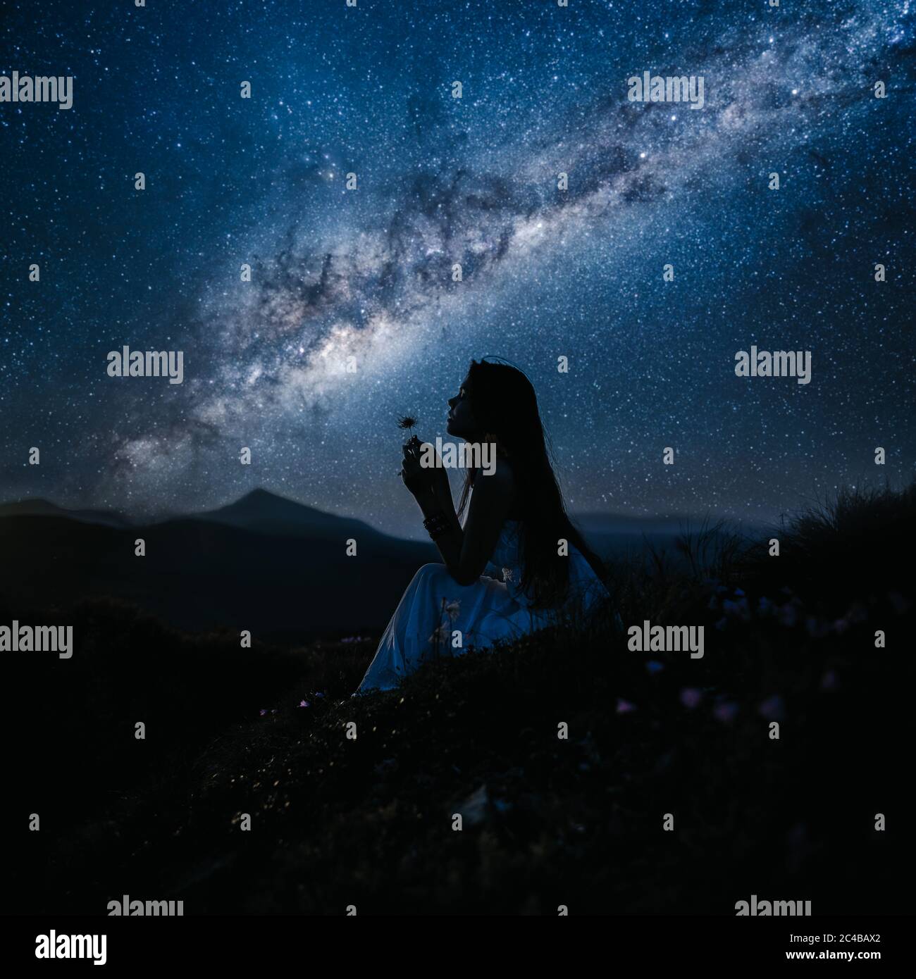 Woman looking at beautiful milky way. Beautiful woman in a long white dress in the mountains. Girl sitting on a rock. Milky Way at mountains. Night Stock Photo