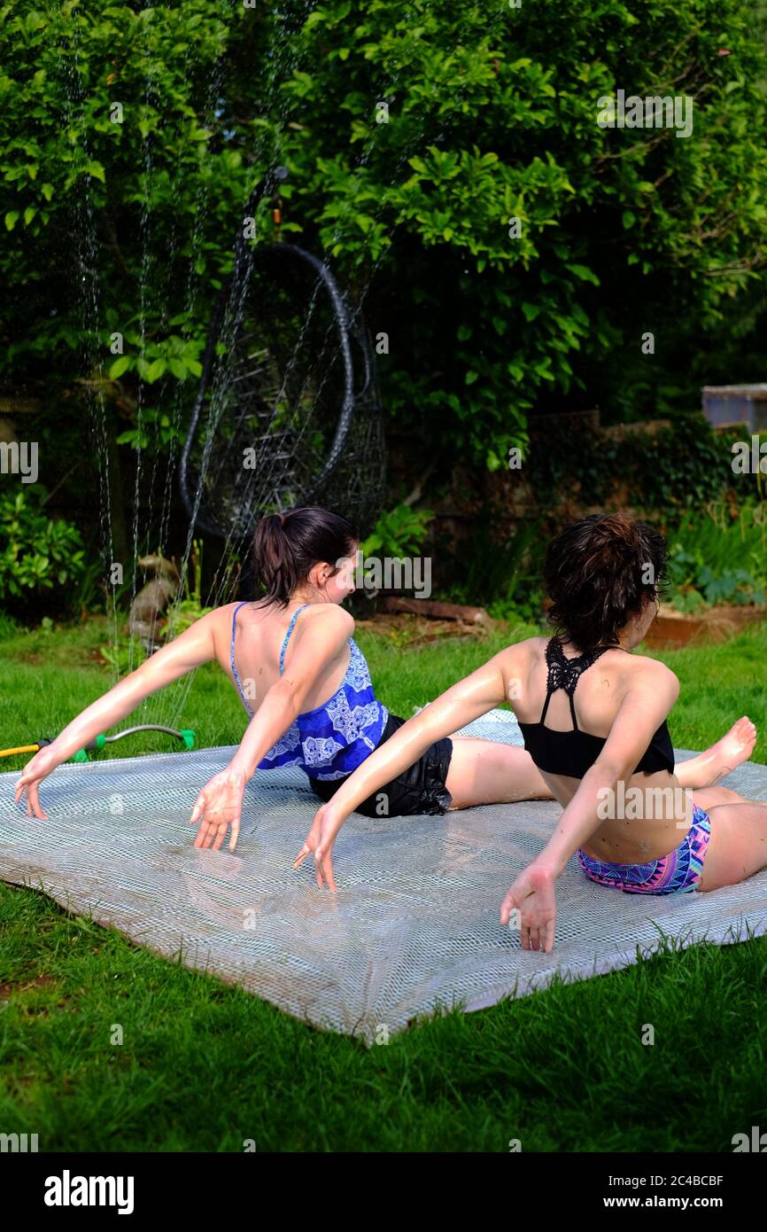Children cooling off during heatwave playing in the garden with sprinkler on plastic sheet slip slide. 25th June 2020 Devon UK Stock Photo