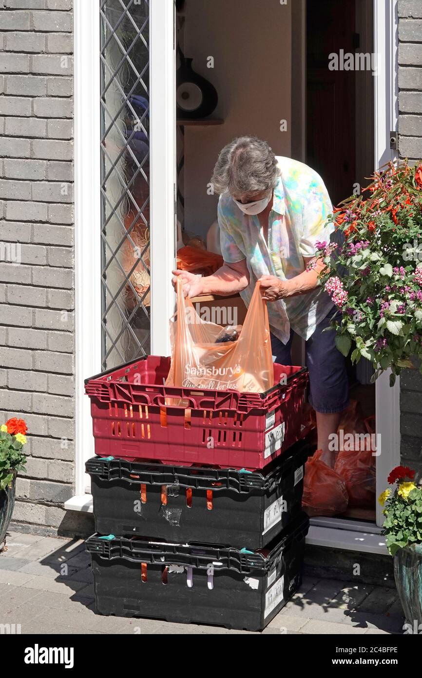 Senior woman pensioner housewife in coronavirus covid 19 lockdown in mask at front door lifts Sainsburys home food grocery delivery shopping bag UK Stock Photo