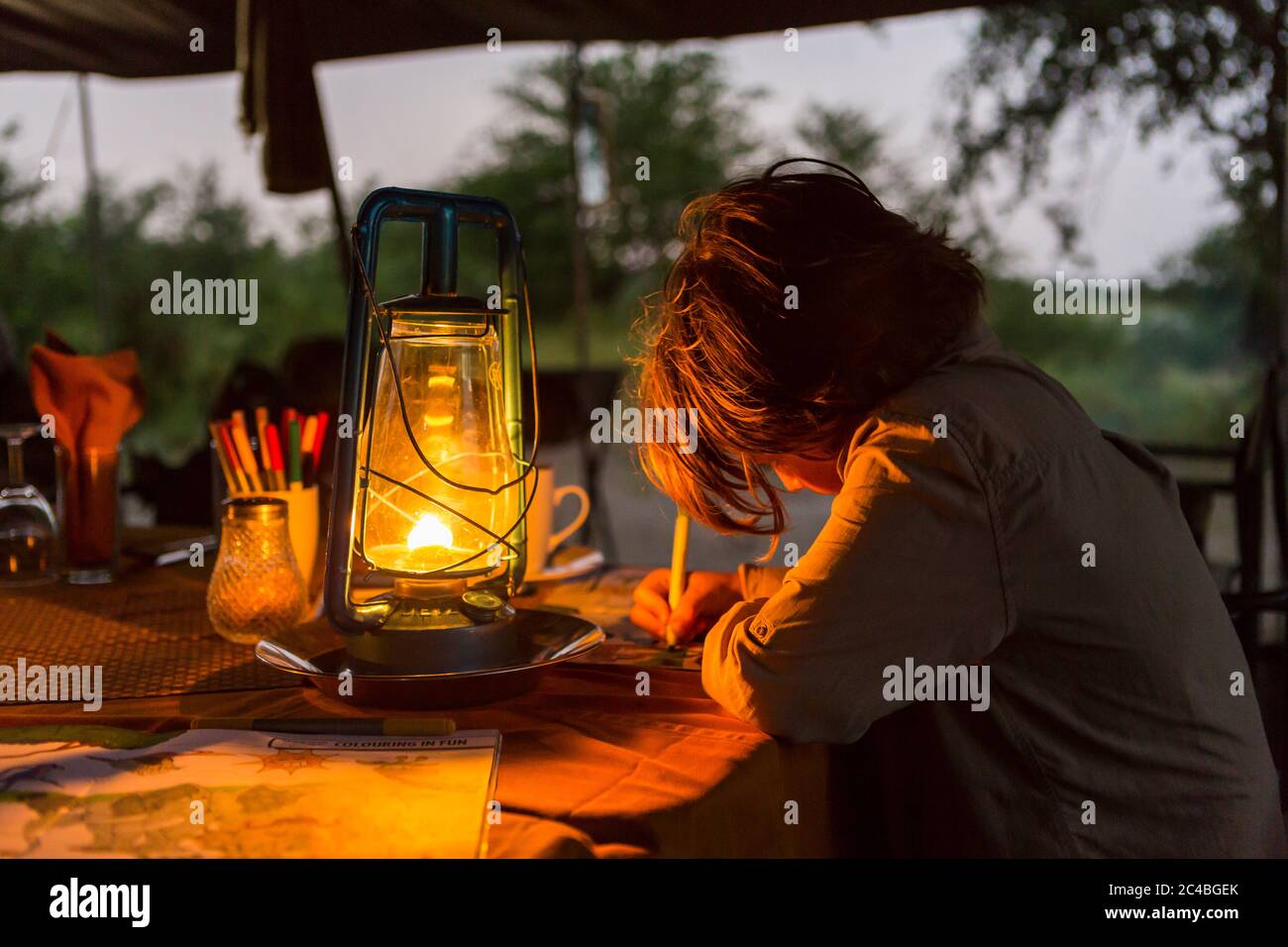 Six year old boy writing a journal by lantern light in a tented camp. Stock Photo