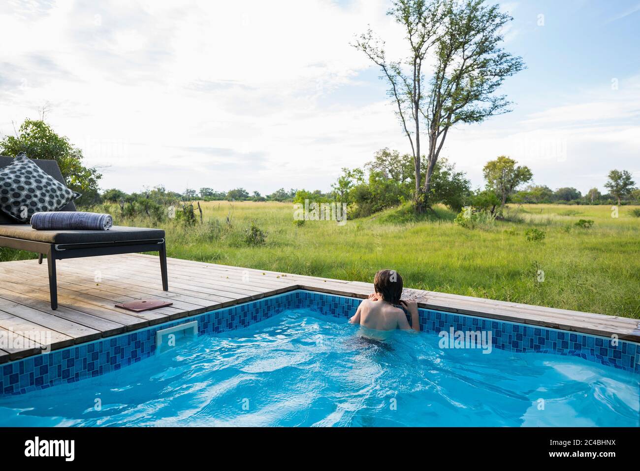 A boy in a swimming pool looking out at the landscape around a safari camp Stock Photo
