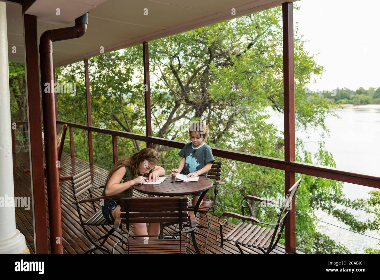 Two children drawing together at a table on a terrace overlooking a wide river. Stock Photo