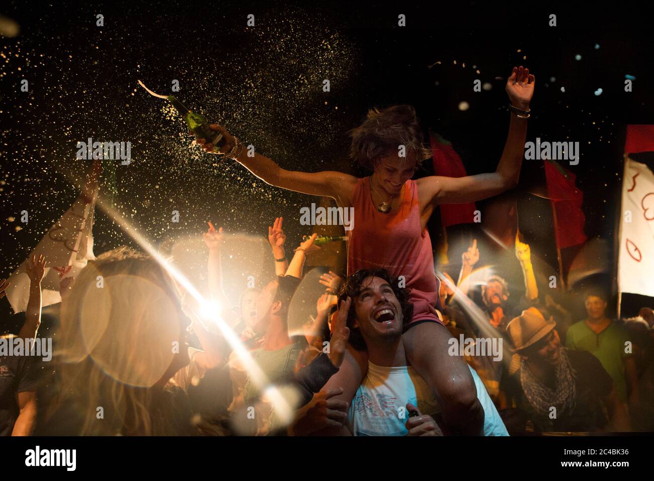 Revellers at an open air concert, smiling man carrying woman on his shoulders, arms outstretched, holding beer bottle. Stock Photo