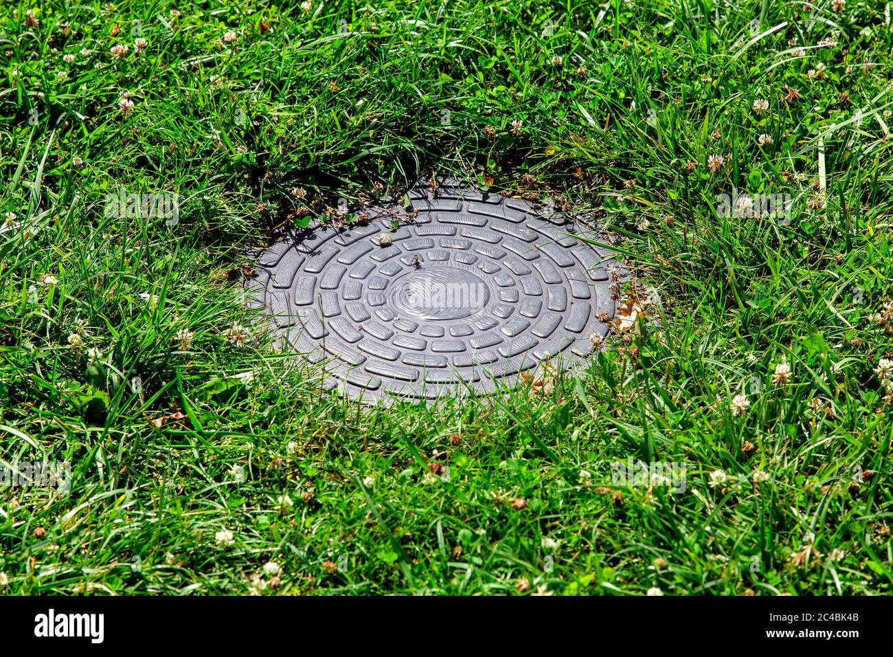 round sewer manhole with a pattern on a green lawn on a sunny summer day. Stock Photo