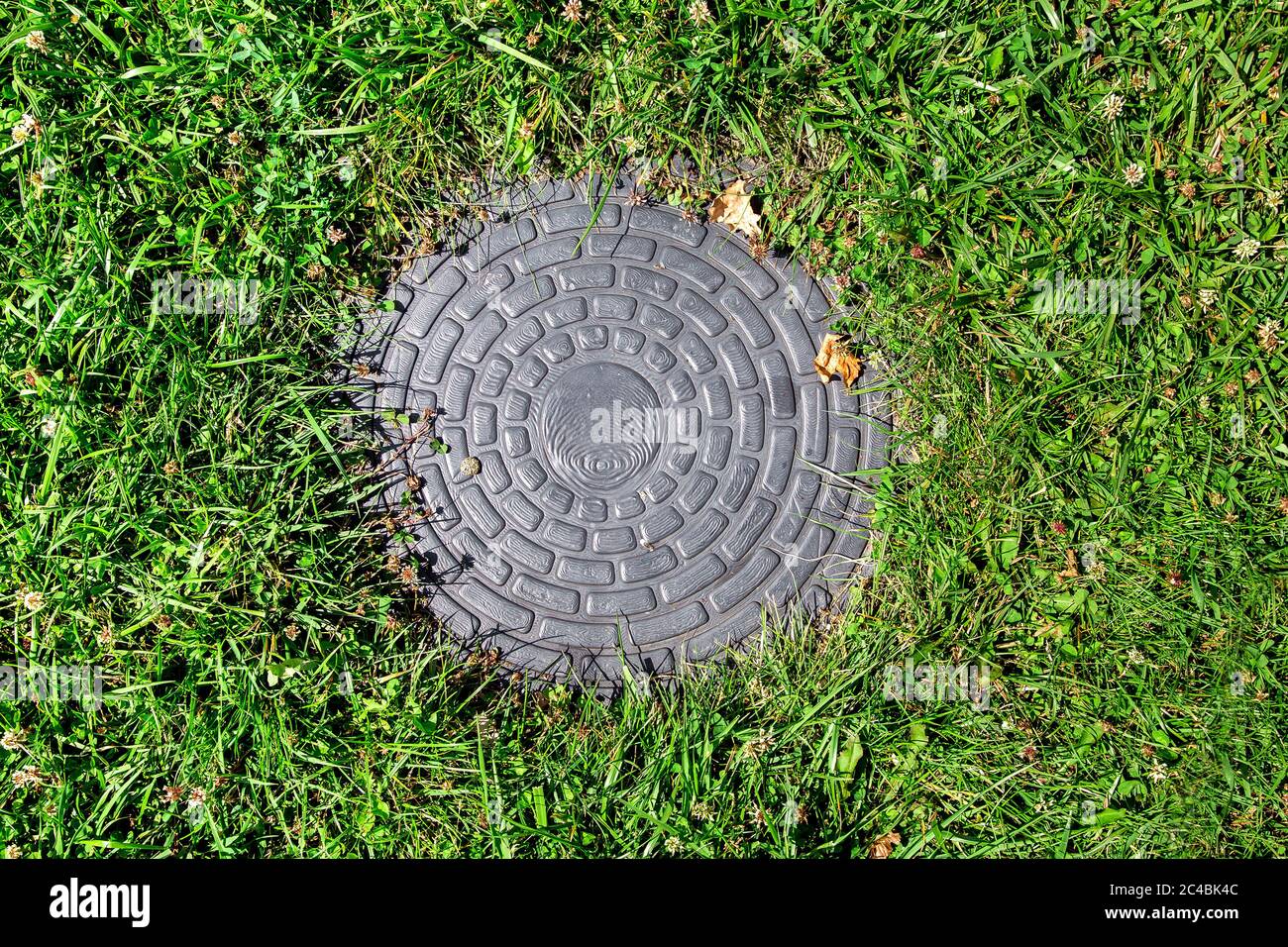 manhole cover in a clearing in green grass on a sunny summer day, top view. Stock Photo
