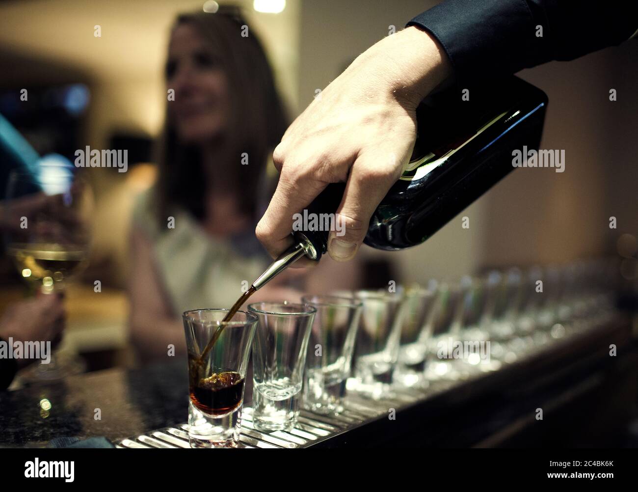 https://c8.alamy.com/comp/2C4BK6K/close-up-of-bartender-pouring-drinks-from-bottle-into-a-row-of-shot-glasses-standing-on-bar-counter-woman-sitting-in-background-2C4BK6K.jpg