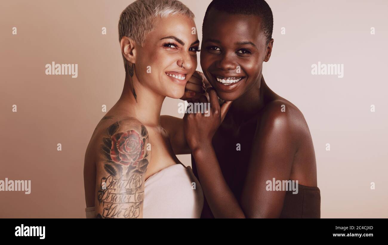 Two beautiful women standing together against beige background. Female models of different ethnicities with short hairstyle looking at camera and smil Stock Photo