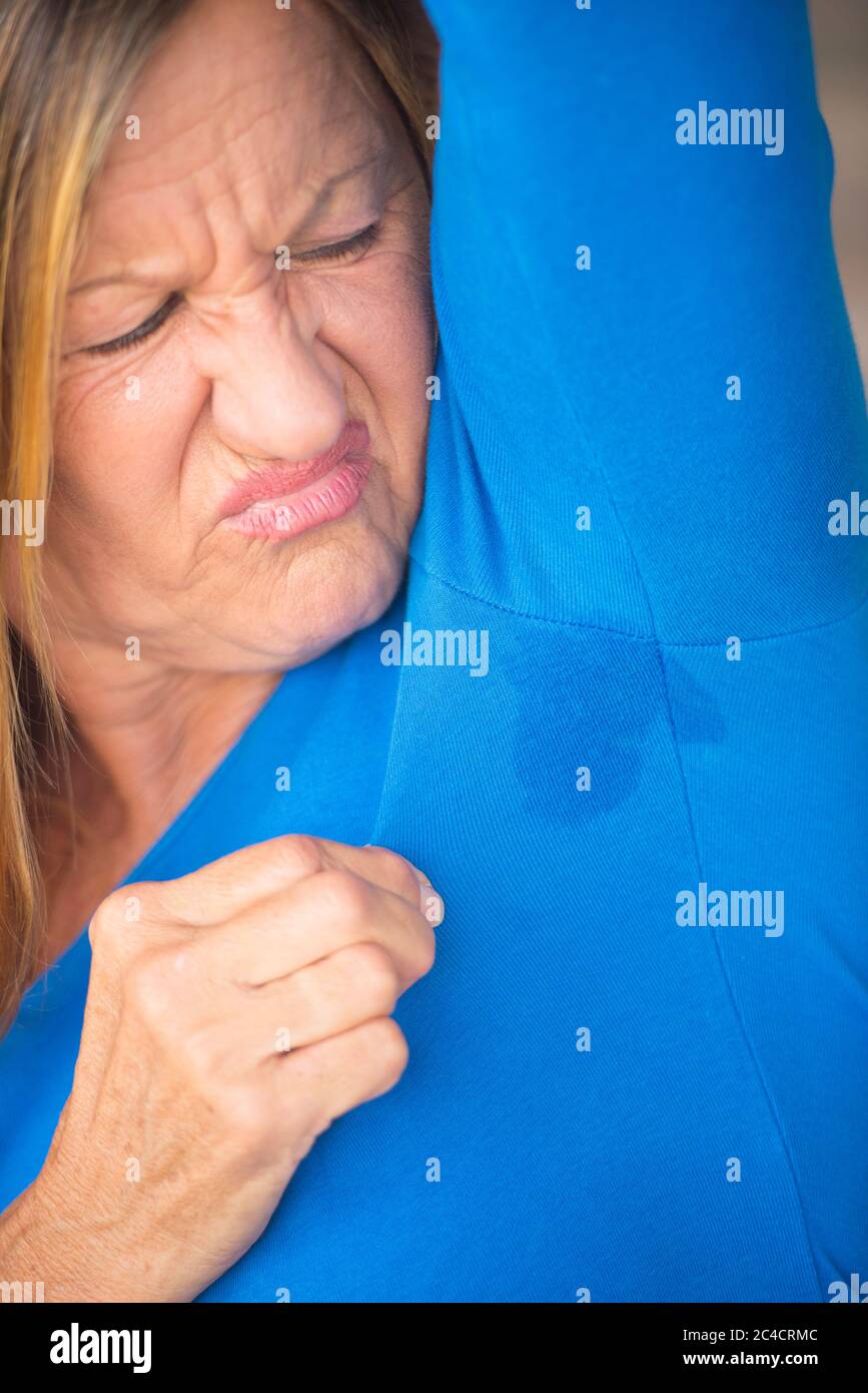 Portrait angry mature woman with sweat perspiration under arm with wet moisture spot on blue shirt, blurred background, copy space. Stock Photo