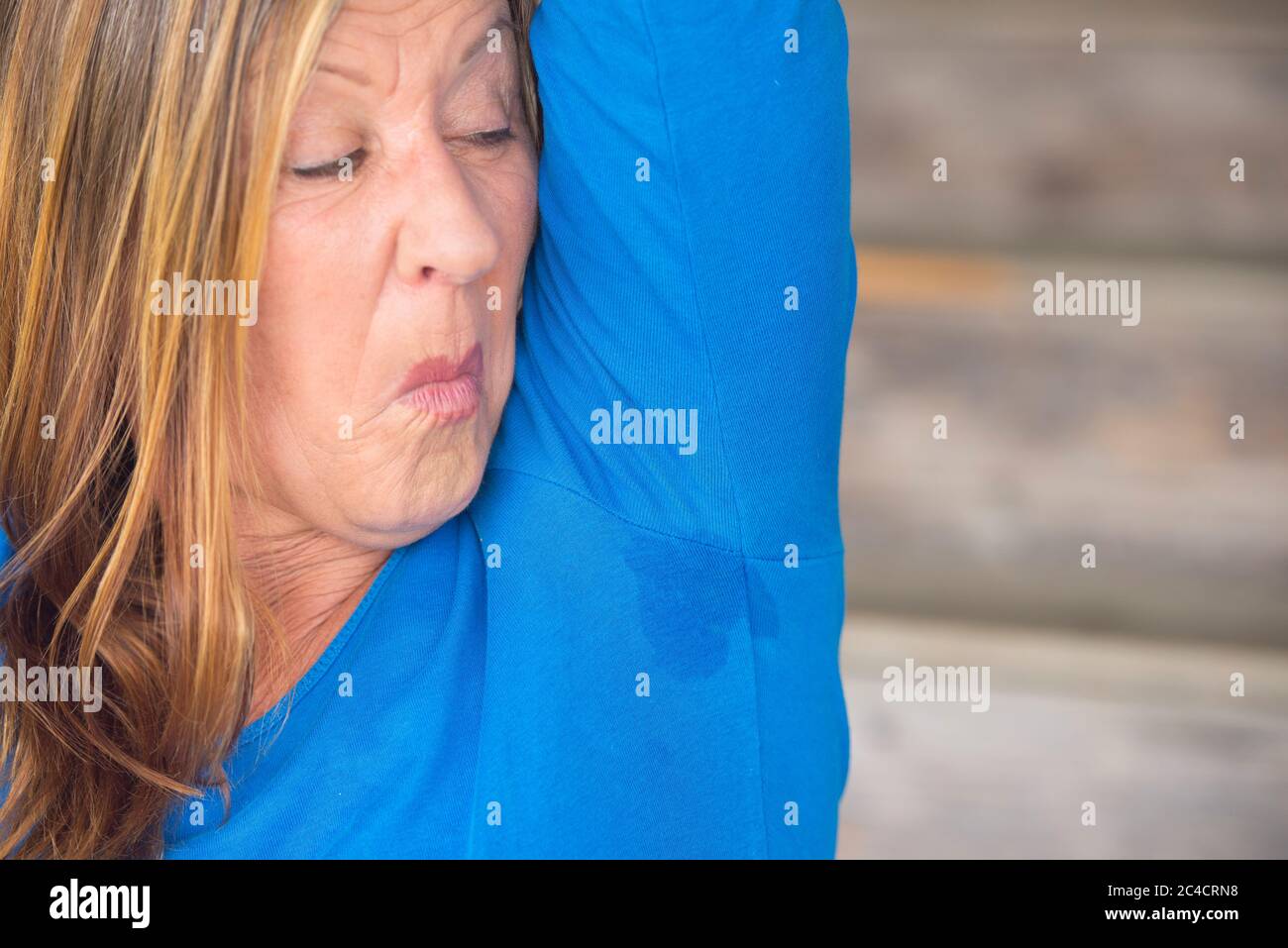 Portrait mature woman with sweat perspiration under arm with smelly wet moisture spot on blue shirt, blurred background, copy space. Stock Photo