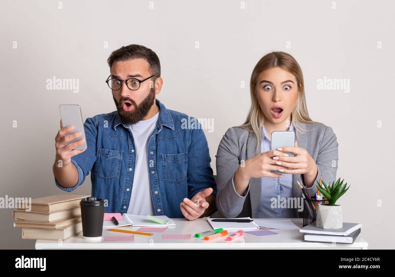Shocking news at work. Colleagues guy and girl are sitting near, with open mouths and looking at smartphones Stock Photo