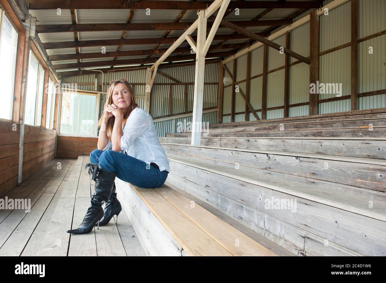 Portrait attractive mature woman sitting thoughtful, sad and concerned on bench, wearing jeans, blouse and stiletto boots, copy space. Stock Photo
