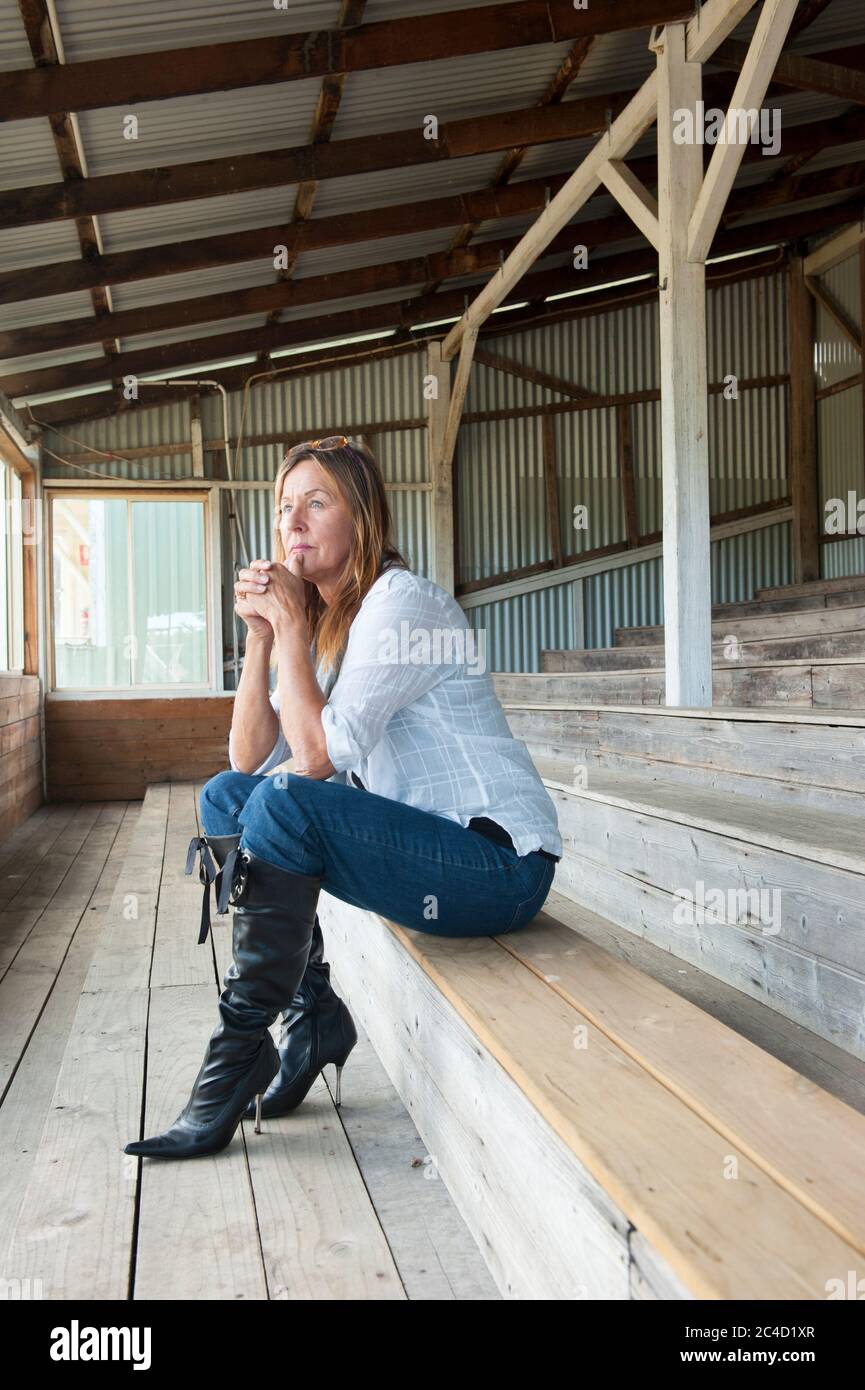 Portrait attractive mature woman sitting thoughtful, sad and concerned on bench, wearing jeans, blouse and stiletto boots. Stock Photo