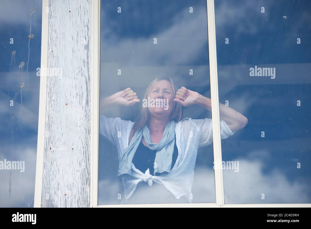 Stressed mature woman crying in despair behind window inside house or home, depressed, sad, lonely, unhappy expression, copy space. Stock Photo