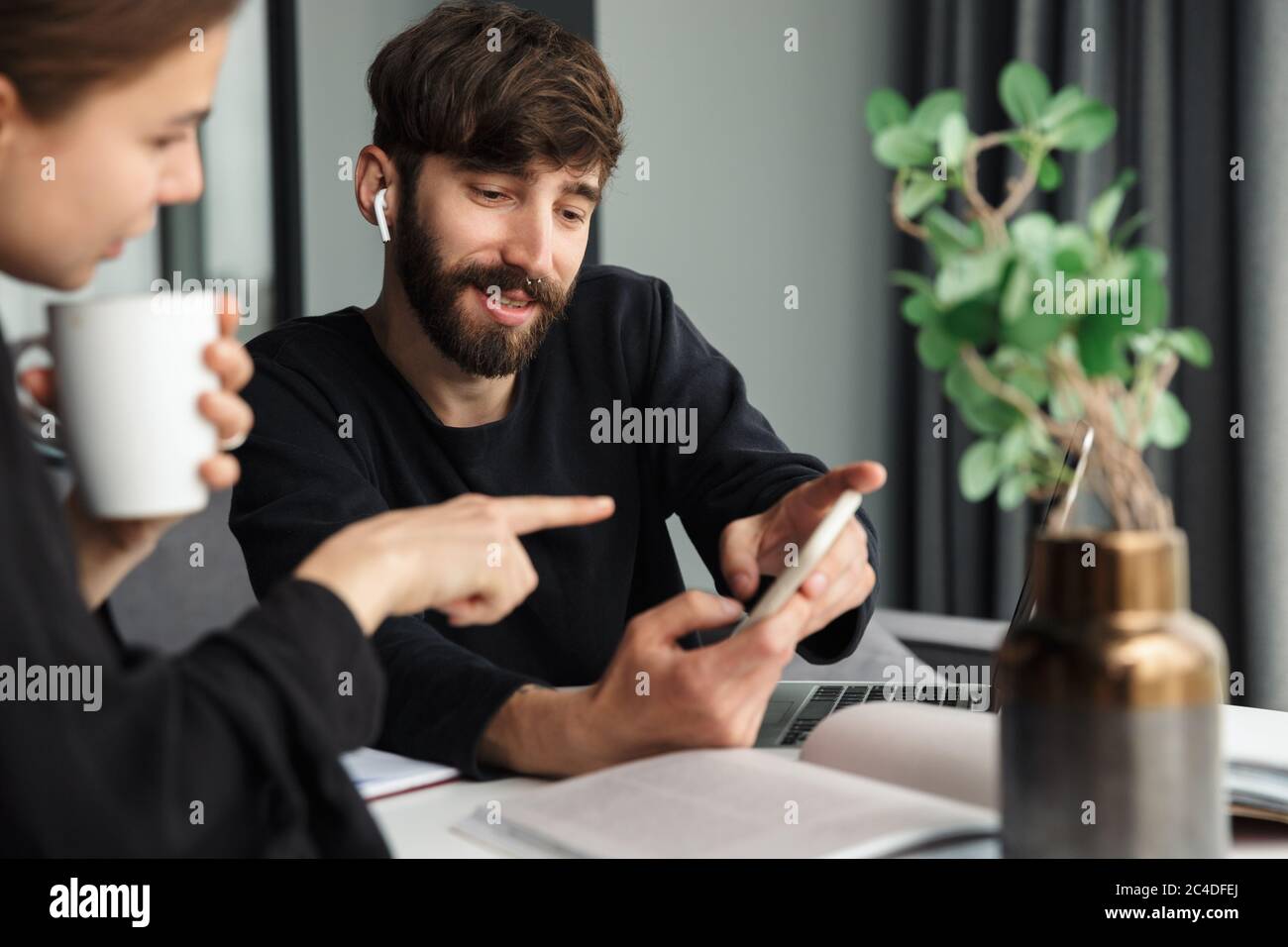 Image of young joyful colleagues discussing project and using cellphone while working at table in cozy room Stock Photo